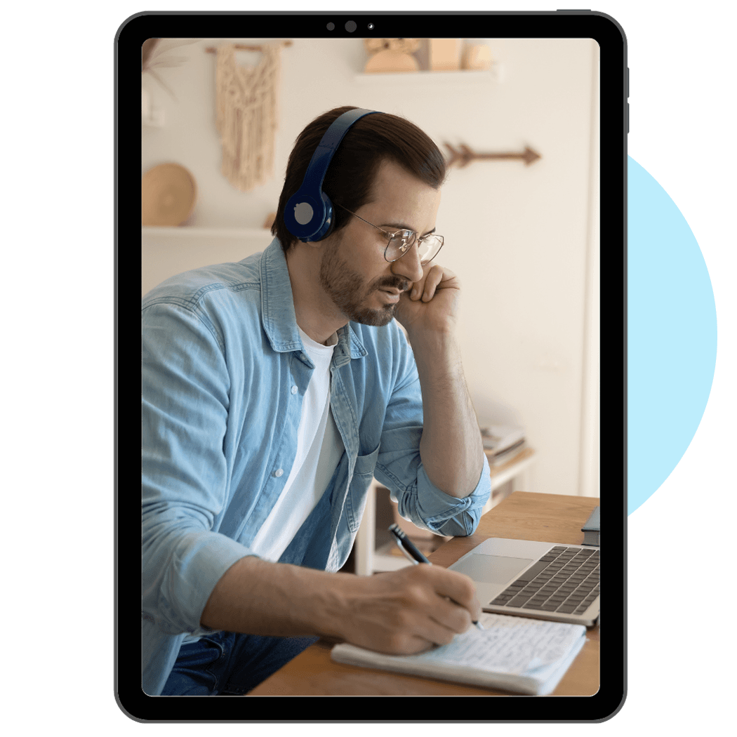 Man with headphones writing in a notebook while using a laptop.