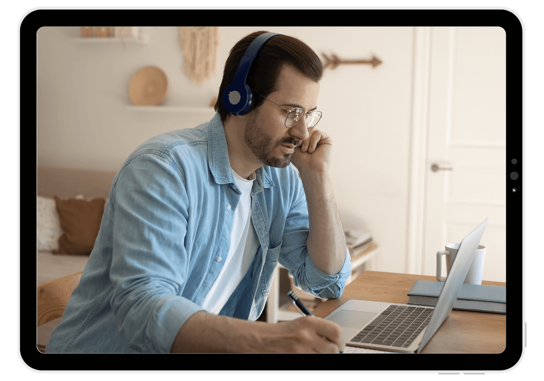 Man with headphones writing in a notebook while using a laptop.